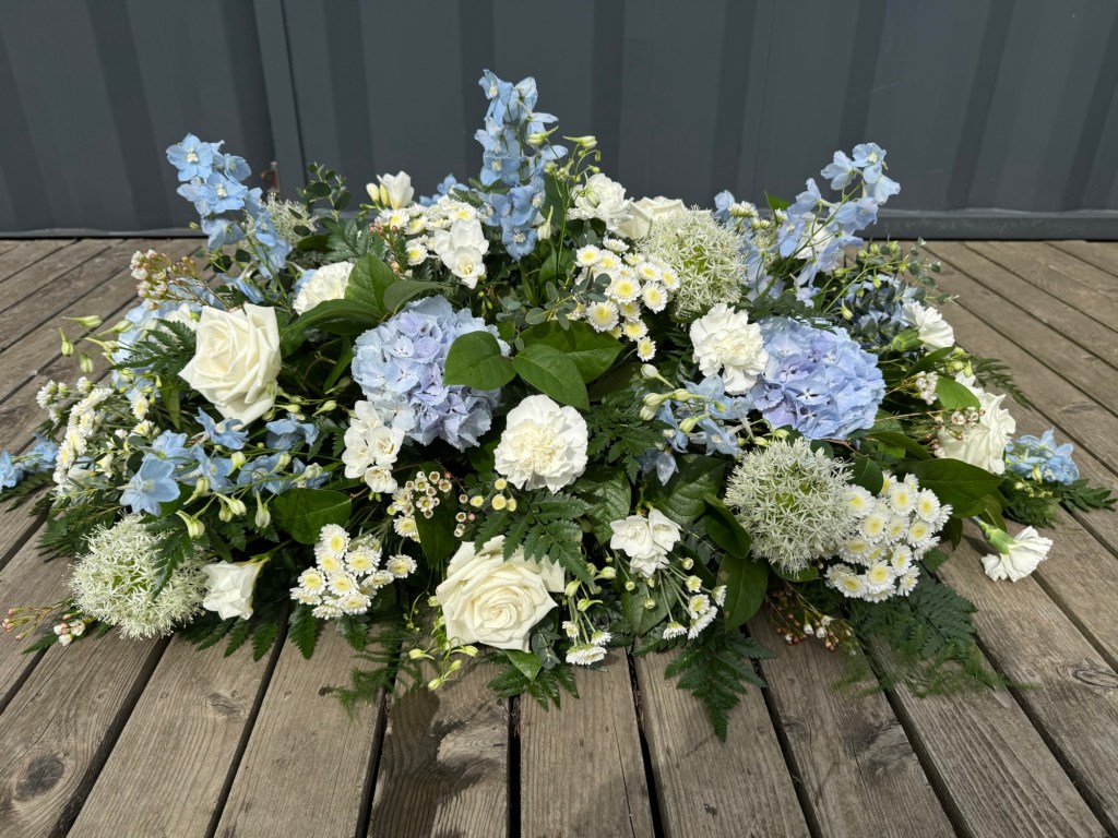 White and pale blue funeral casket spray, made with hydrangea, delphiniums, allium, carnations, freesia and roses.